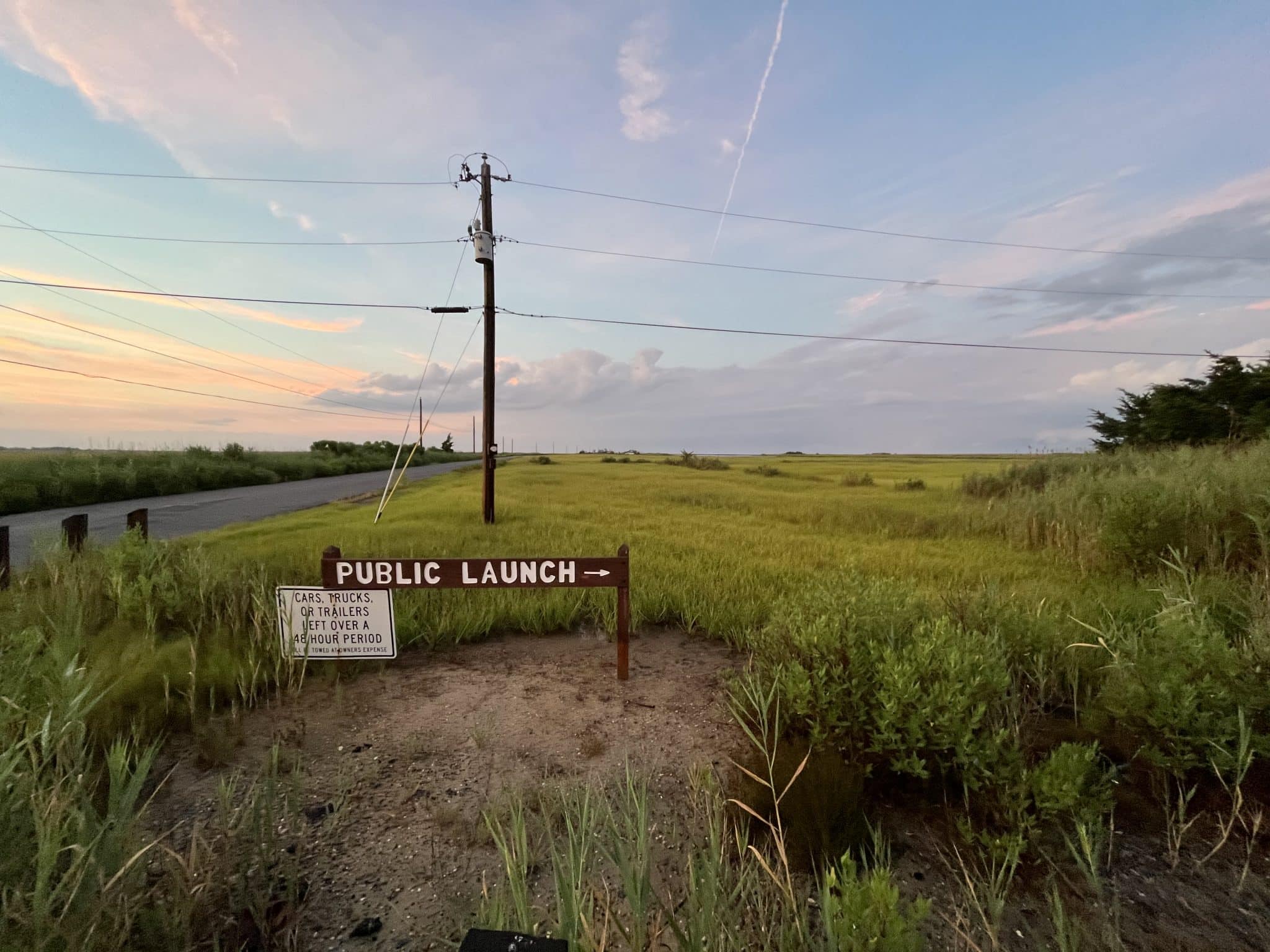 Two roadside signs reading "Public Launch" and "Cars, trucks or trailers left over a 48 hour period will be towed at owner's expense." The background is an expanse of marsh and a pink dusk sky.