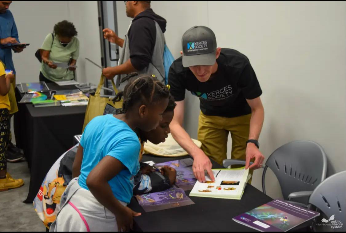 Two children lean over a table with brochures, while a man with a hat that says Xerces Society points to the photo of a firefly in a book.