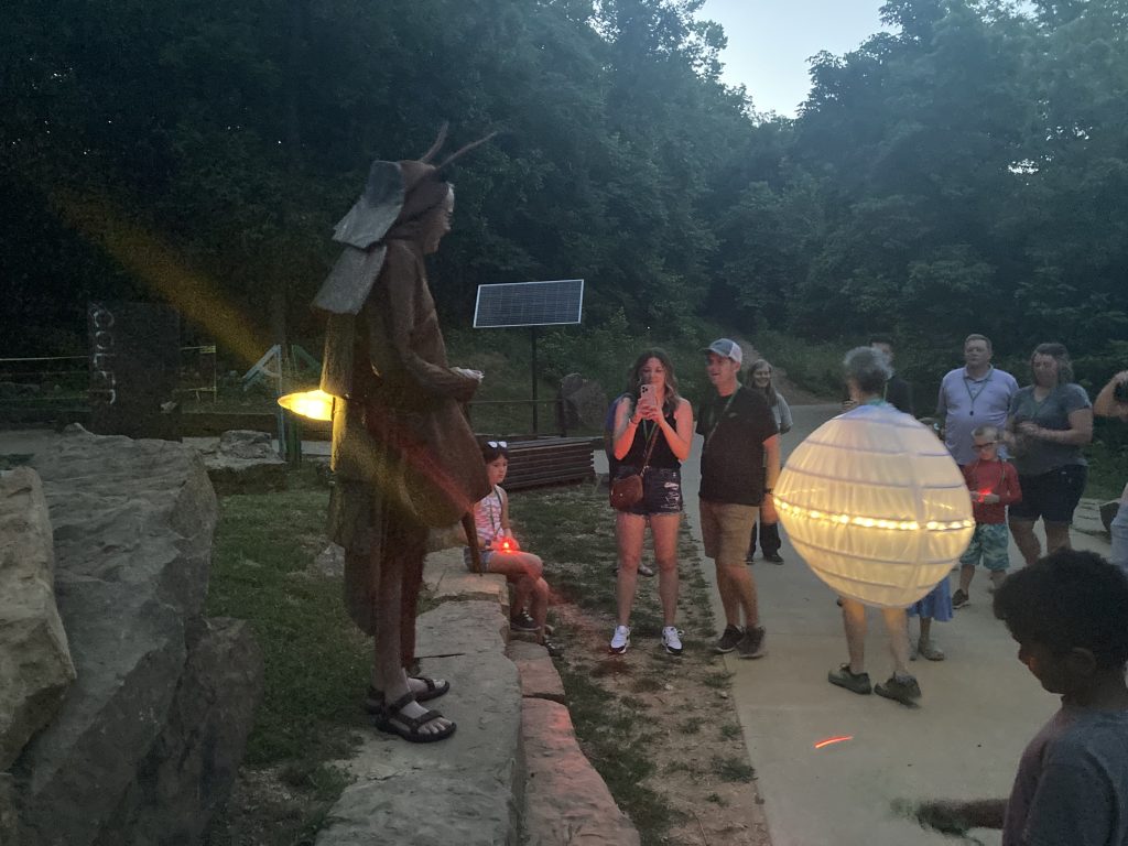 A person dressed as a firefly larva speaks to a small group of people at a firefly viewing event. In the crowd walks another person dressed as a glowing firefly egg.