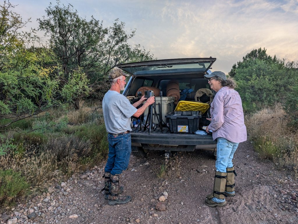 Image shows two biologists standing at the back of an open truck parked on a dirt road. The truck is filled with camping and field gear. On the open tailgate, one biologist is fiddling with some camera settings on a GoPro setup while the other talks to them.