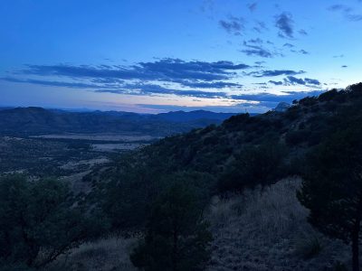 Looking out from a rocky hillside onto a darkening valley below at twilight. An observatory sits in the distance.